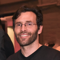 A white man with beard and glasses wearing black sweater smiles at camera