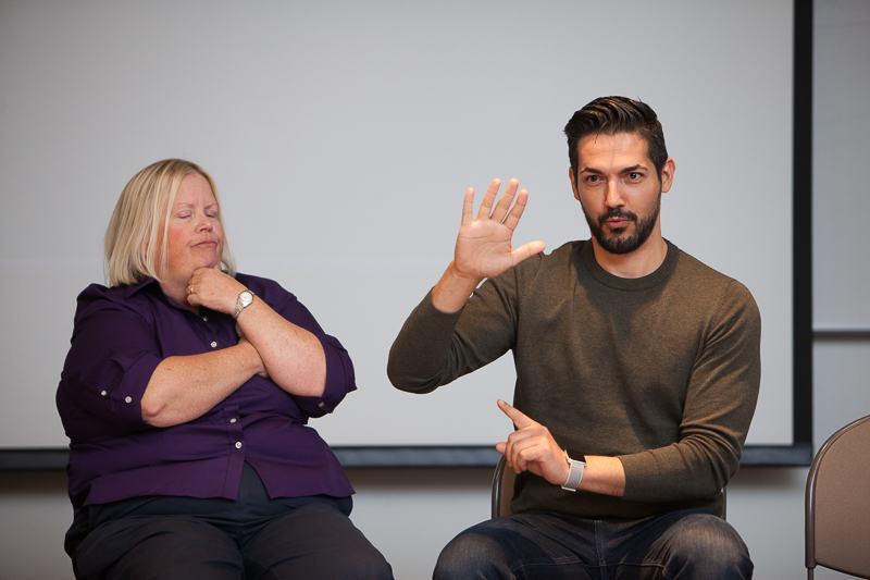 A white woman wearing a purple shirt looks on as a Latino man with a beard wearing a brown shirt signs