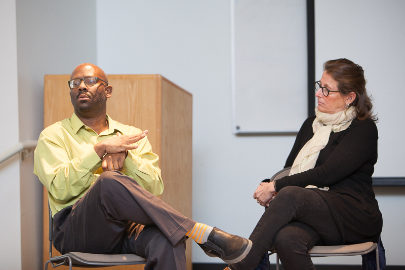 A black man with glasses and bald head sits as part of panel. A white woman with black glasses wearing a black shirt and white scarf looks at him.
