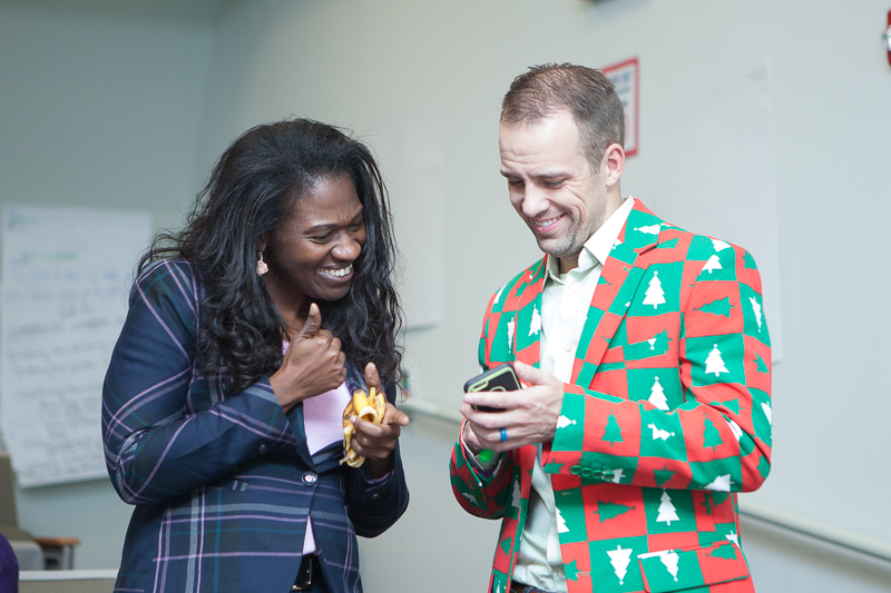 A black woman with long black hair smiles and looks at white man wearing red, green, and white sportcoat who is showing her something on his phone.