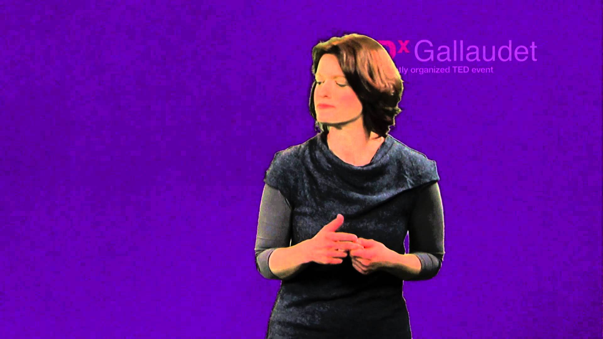 A white woman with brown hair wearing a blue shirt clasps her hands in front of a purple background