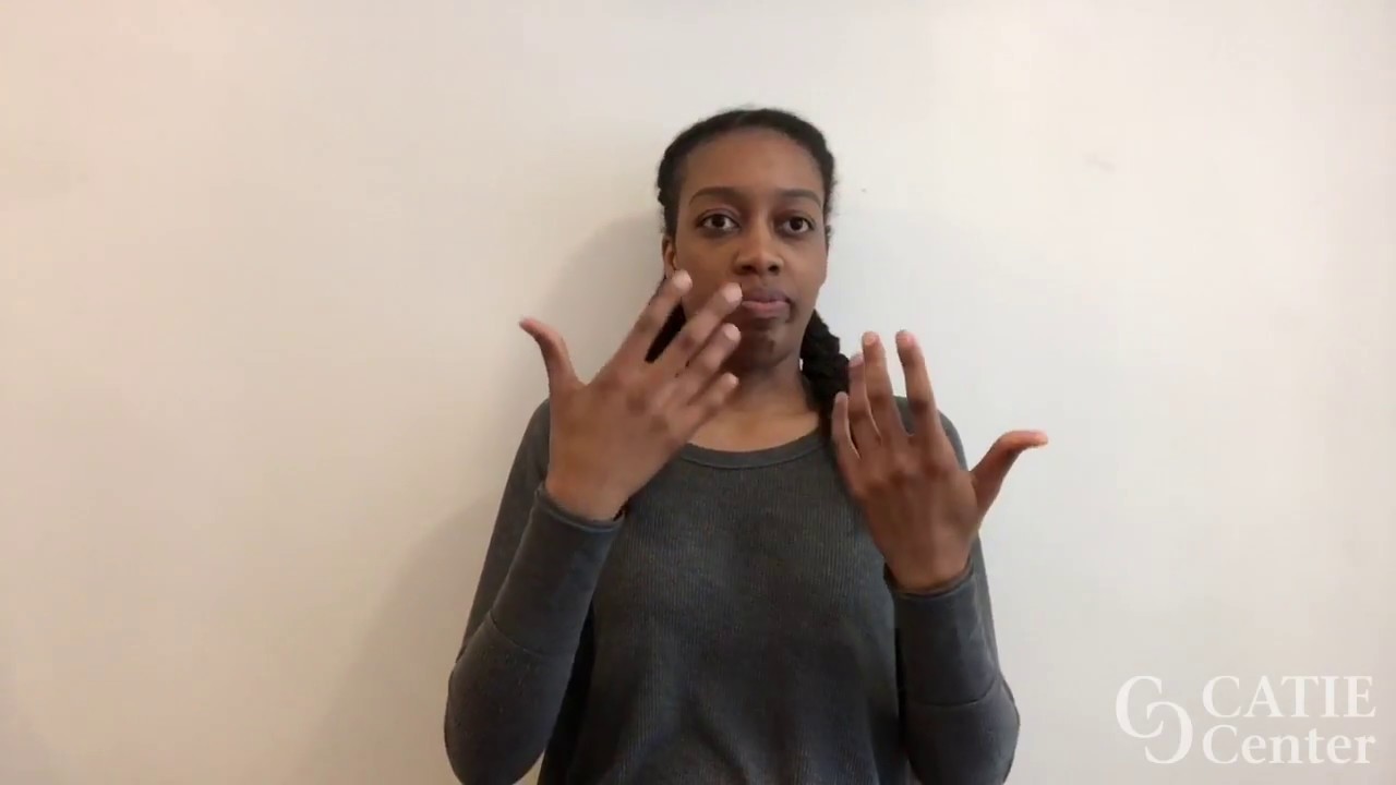A black woman wearing grey long-sleeve shirt holders her hands up with palms facing toward her face ready to sign