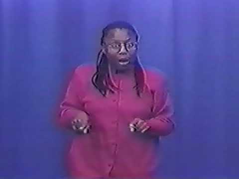 A black woman with braids and glasses wearing a pink top stands in front of a blue background and narrates an ASL text