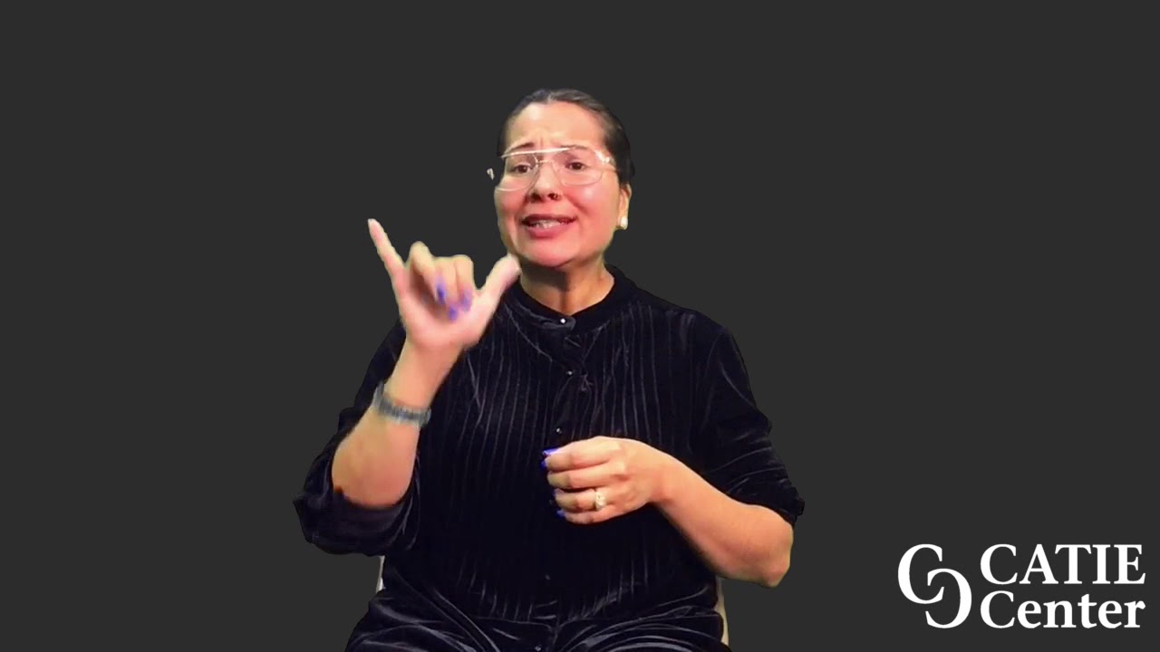 A woman with fair skin is wearing a black blouse with light vertical stripes down the front.  She is wearing wire-rimmed glasses and has her dark hair pulled back.  She sits in front of a dark grey background and narrates the entire story.  In the lower right corner of the video screen is a white CATIE Center logo.