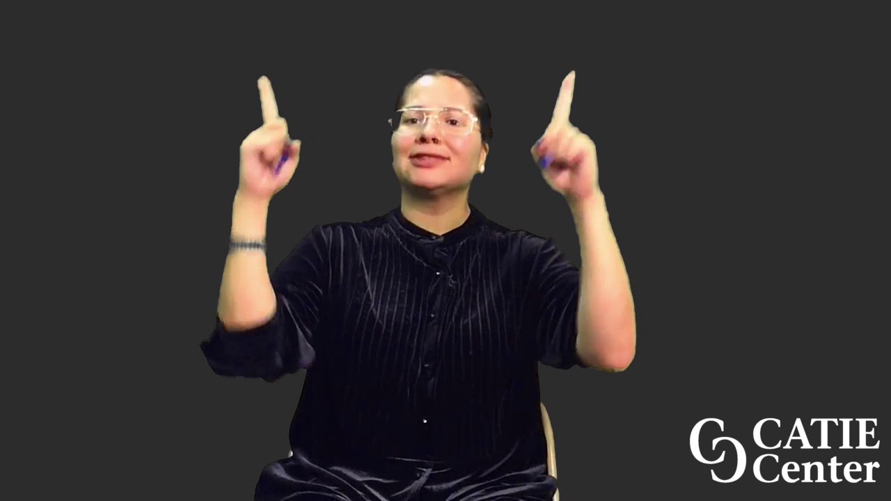 A woman with dark black hair pulled back and glasses wearing a black velour shirt sits and holds her two index fingers up.