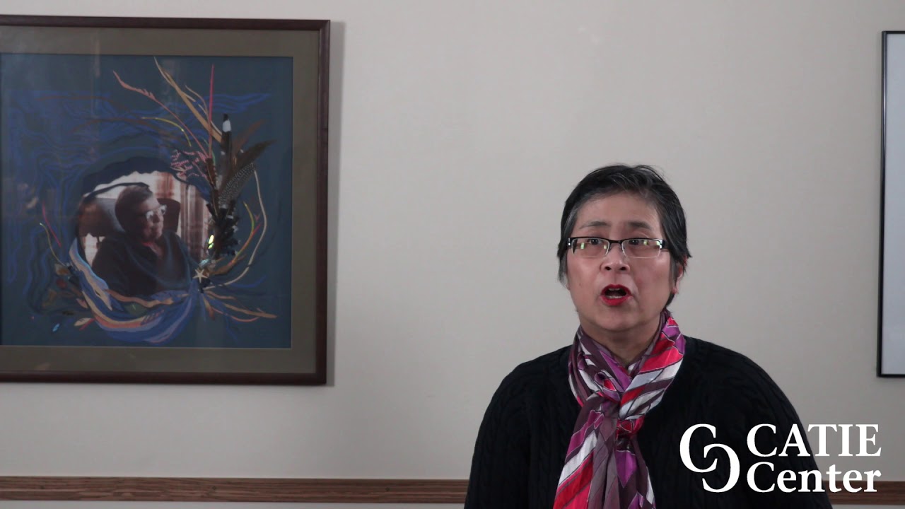 A Chinese-American woman with short black hair and glasses wearing a colorful scarf speaks in front of a tan wall.