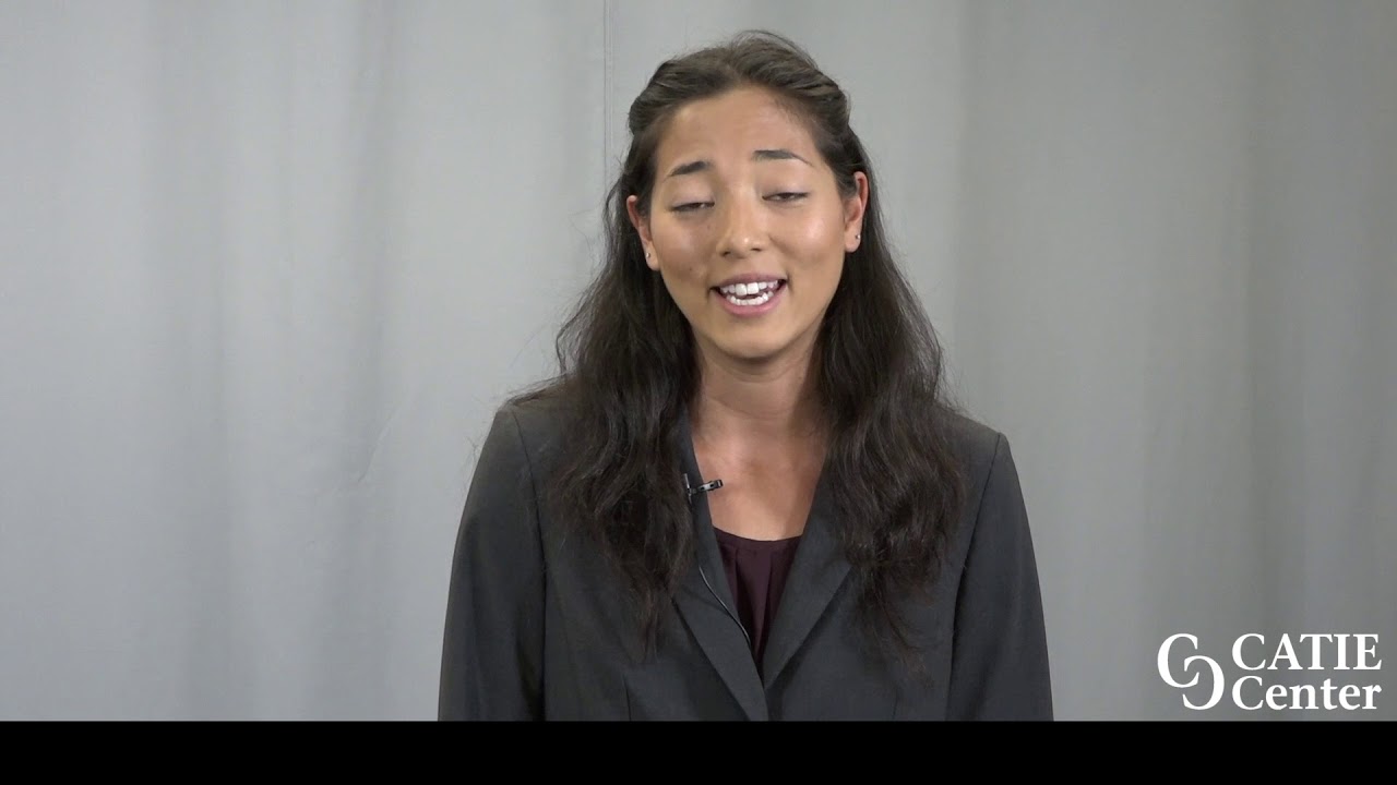 A woman with light brown skin and dark brown long hair wearing a grey suit coat stands in front of a light grey background and speaks.