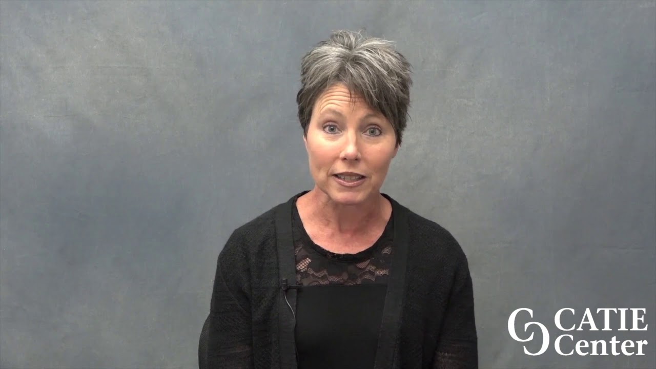 A white woman with short salt and pepper hair wears a black shirt in front of a grey background speaking in English. The CATIE Center logo is in the lower right corner of the screen.