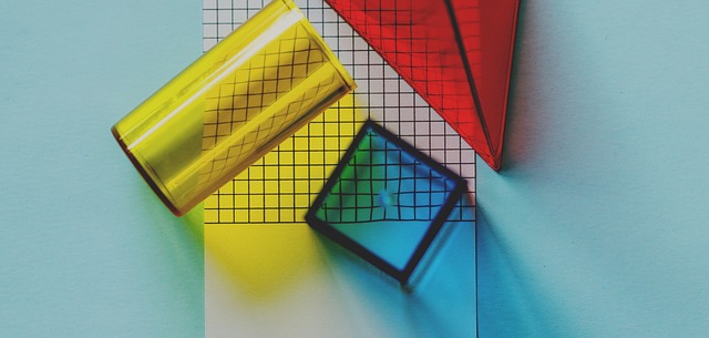 a Yellow cylinder, blue cube and red pyramid sit on top a piece of white graph paper