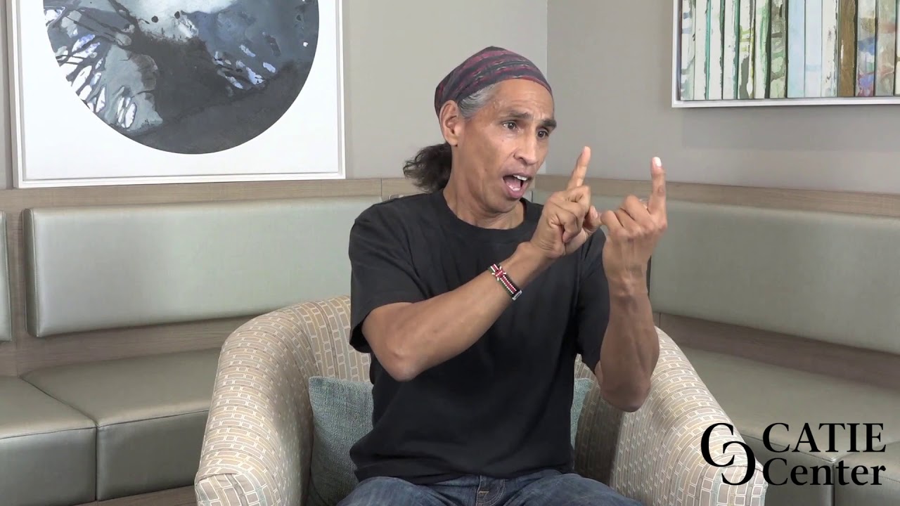 a brown-skinned man sits in a white chair wears a black short sleeved shirt and a bandana on his head - signing "Meet"