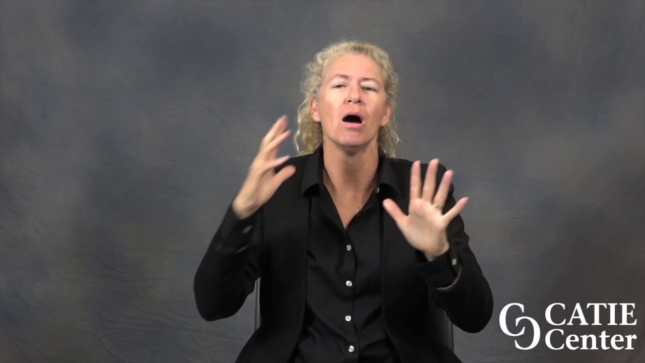 A white woman with blonde hair pulled back in a pony tail wearing a black, button down shirt and a black coat signs in front of a blue/grey background. The CATIE Center logo is in the lower right corner of the video screen