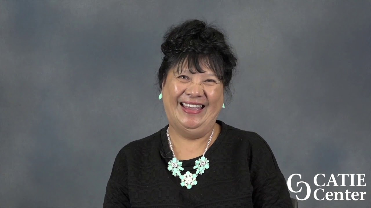 A Filipino woman with black hair pulled up wearing a white necklace and black blouse speaks in front of a blue/grey background. The CATIE Center logo is in the lower right corner of the video screen