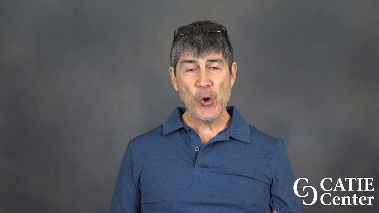 A white man with short brown hair wearing a blue shirt stands in front of a grey background speaking. a White CATIE Center logo is in the lower right of the image.