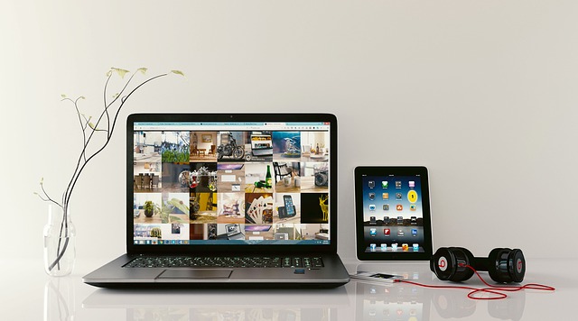 A PC laptop sits on a white counter next to an iPad with headphones plugged into a phone