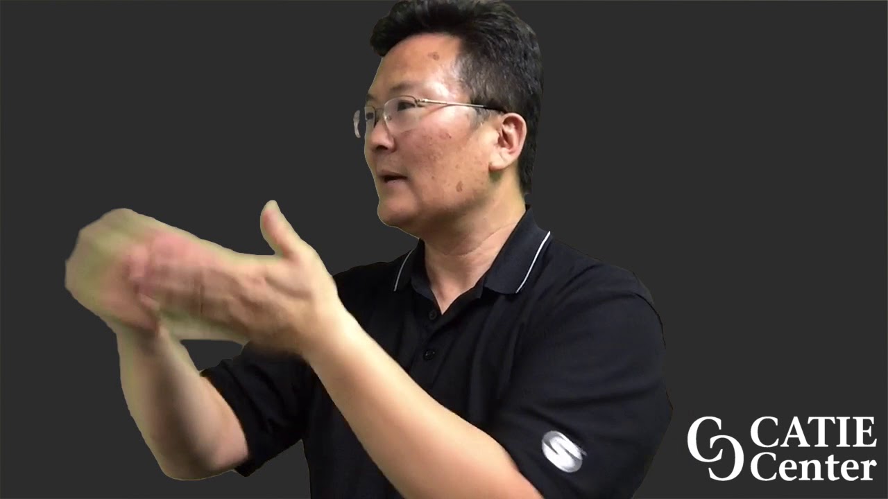 an Asian-American man wearing glasses and a black polo short-sleeve shirt is shown from the waist up. He is standing in front of a dark-grey screen. A white CATIE Center logo is in the lower right corner of the screen. The man narrates the entire video in American Sign Language.