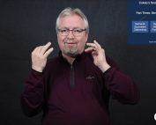 Jimmy Beldon stands in front of a dark grey background.  He is a white man with blond-grey hair parted in the middle wearing black glasses with a goatee.  He is wearing a dark red quarter zip sweatshirt.  In the upper right of the video window, a series of slides shows different aspects of the Cokely model.  The CATIE Center logo is in the lower right of the video window.