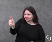 A white woman with dark brown hair wearing a black sweater stands in front of grey background and gives a thumbs-up sign