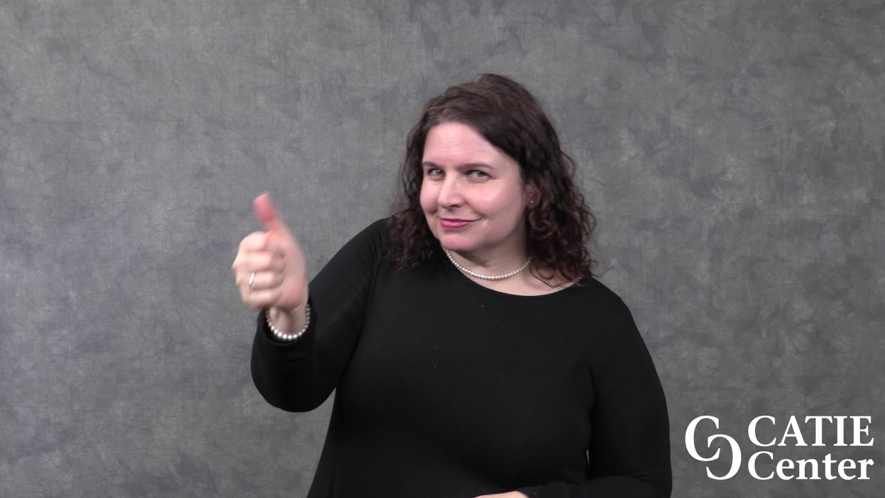 A white woman with dark brown hair wearing a black sweater stands in front of grey background and gives a thumbs-up sign