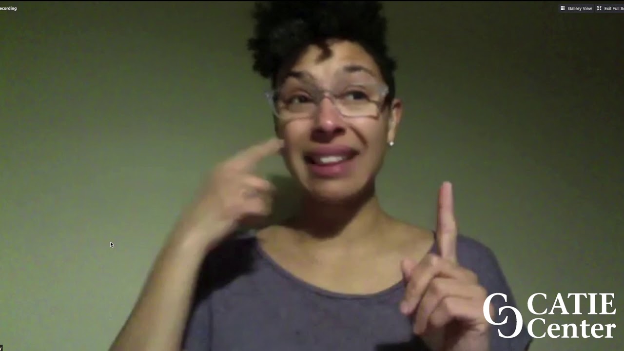 A person presenting as female with brown skin and short curly hair wearing glasses and a purple shirt signs in front of a green wall