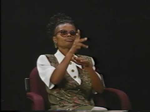 A black woman wearing glasses and a brown vest and white shirt sits in a chair and signs her message