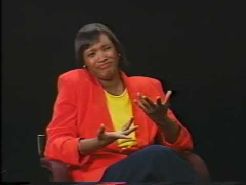 A Black woman wearing a red sportscoat and yellow blouse sits in a chair and signs her message