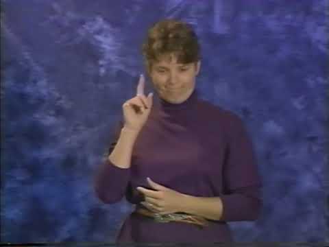 A white woman with short brown hair wearing a purple turtleneck signs in front of a blue background