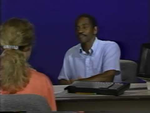 A black man with short hair and moustache sits across a desk from a woman with brown hair. You can only see the back of the woman.