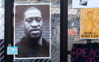 Photo of a black man wearing a black hoodie posted in a bus stop next to a poster with the words Justice for George Floyd